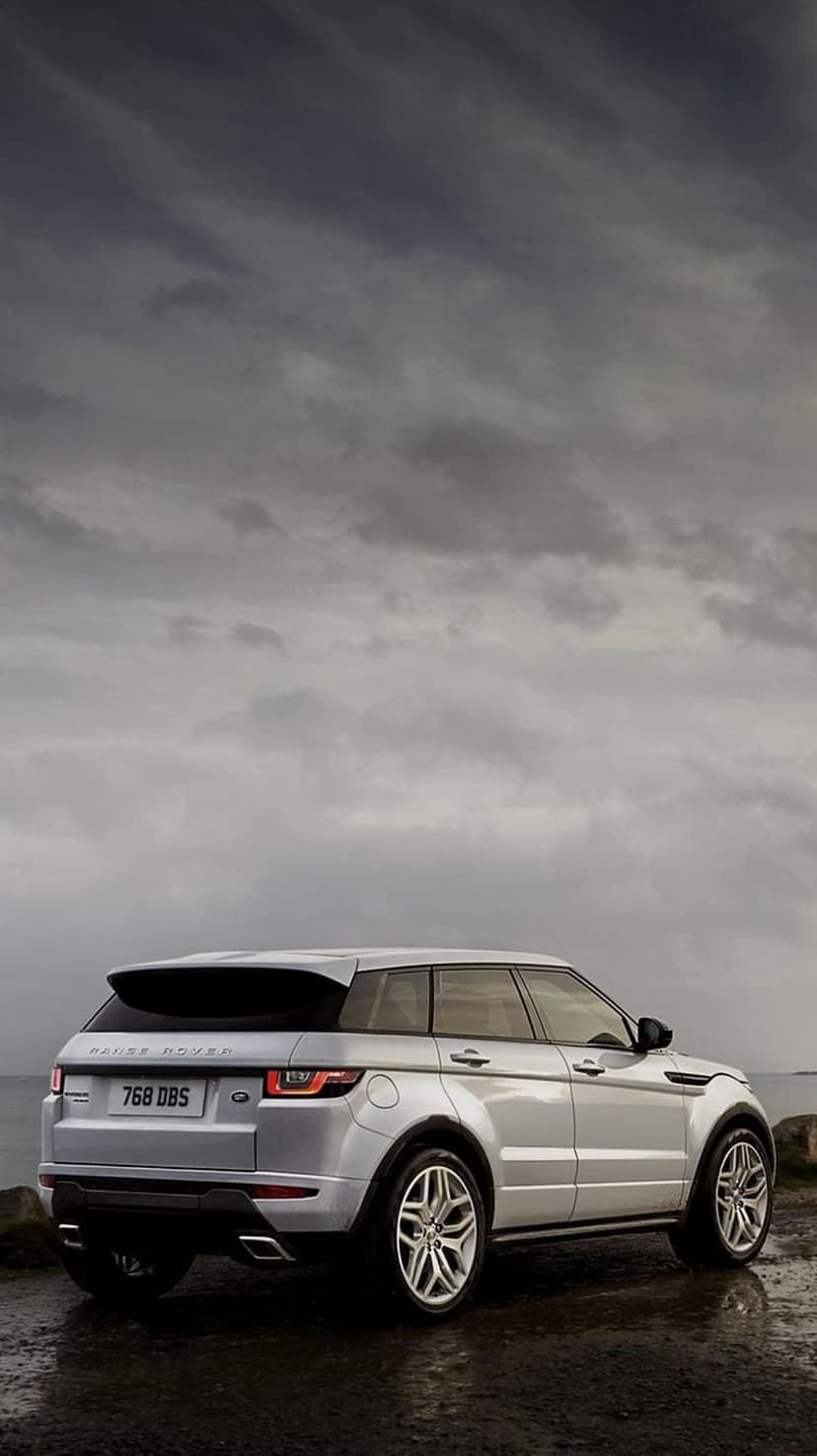 Silver Range Rover Evoque SUV parked on wet pavement beneath a dark, overcast sky showing rear taillights.