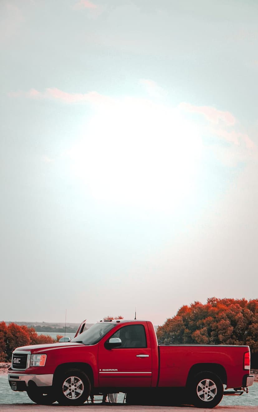 Red GMC Sierra truck parked on a lakeshore during autumn with colorful orange trees and a soft hazy sky.
