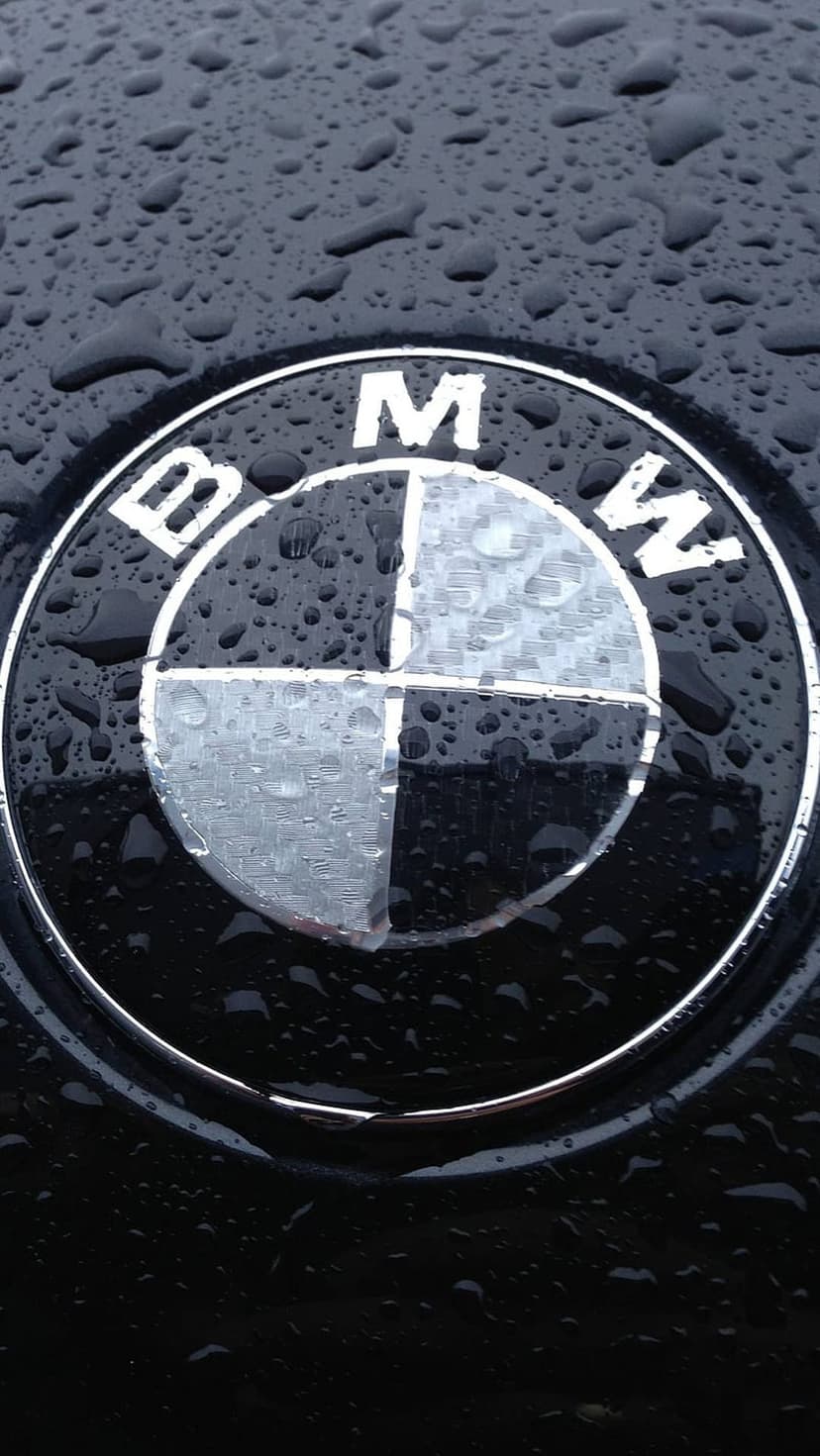 BMW emblem on a black car hood covered in small water droplets during a rain shower with metallic reflections.