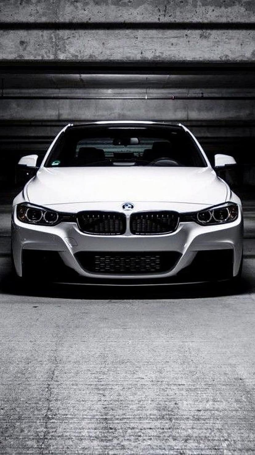 White BMW sedan with glowing headlights parked in a dark, industrial concrete garage with sharp shadows.