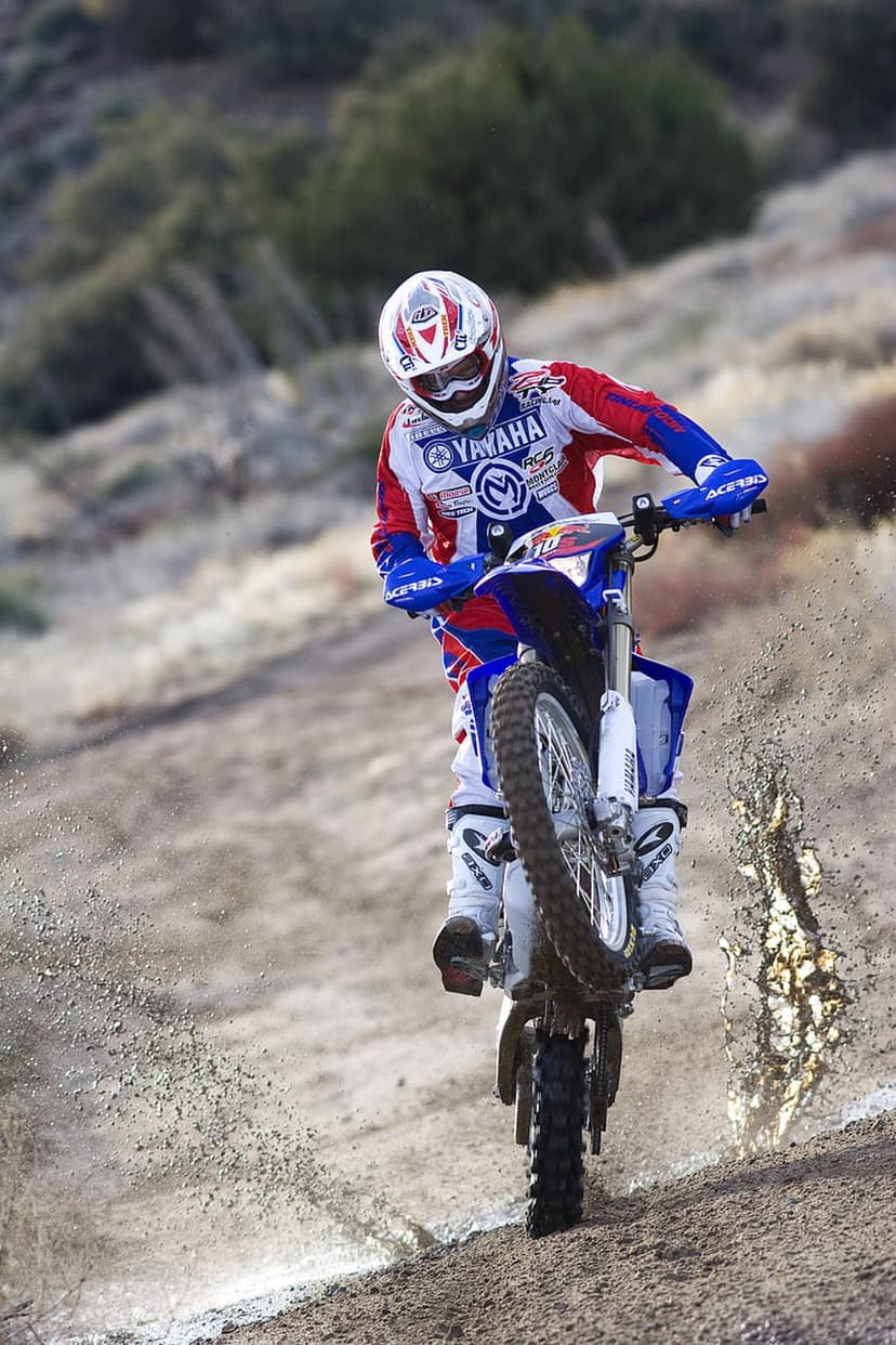 Motocross racer in red and blue gear jumps a Yamaha dirt bike over a sandy hill, kicking up a trail of dust.