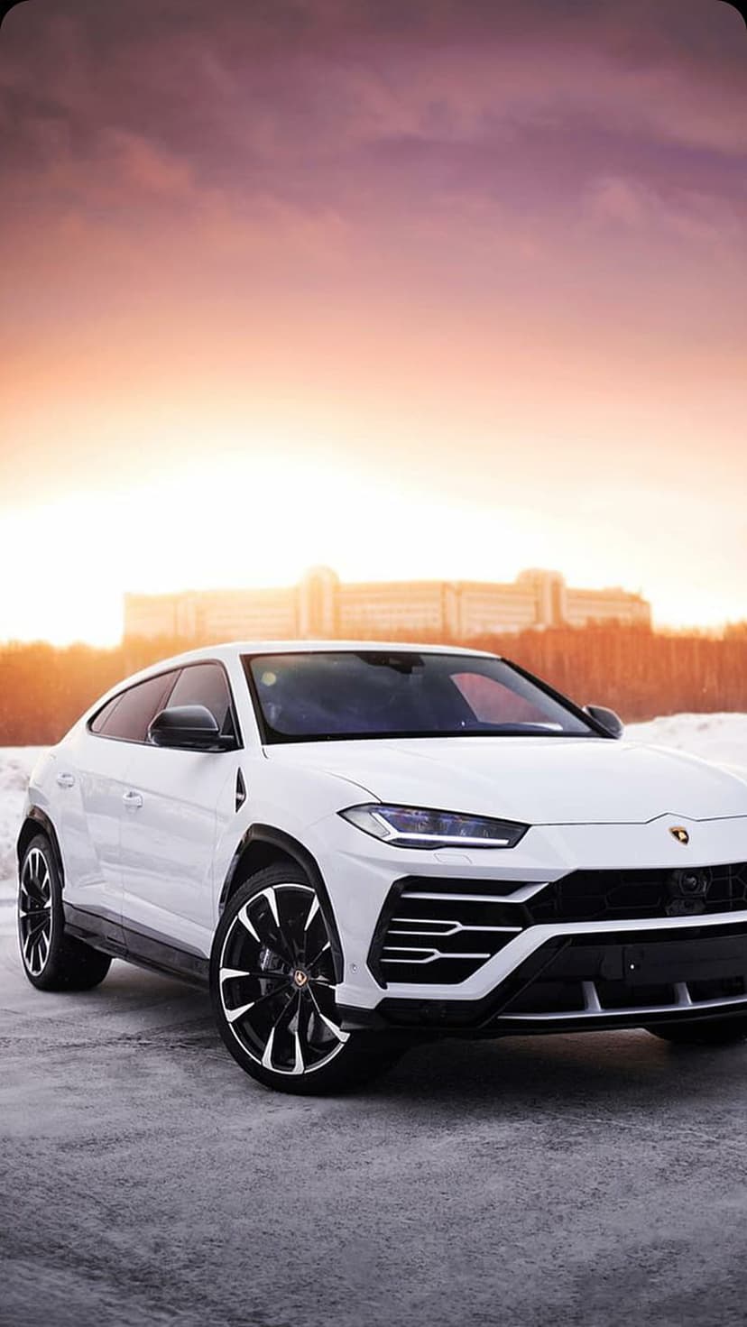 White Lamborghini Urus SUV parked on snow during a vivid orange sunset with a building in the background.