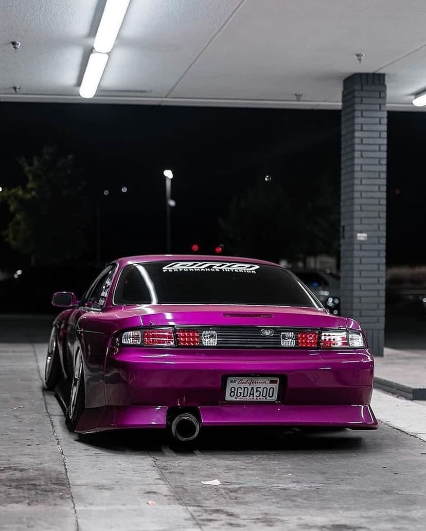 Purple Nissan S14 coupe with custom aero and slammed stance parked under fluorescent lights in a garage.