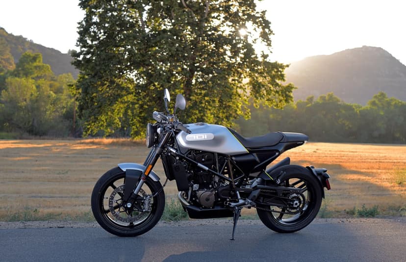 Silver Husqvarna Vitpilen 701 motorcycle parked on an asphalt road at sunset with trees and hills background.