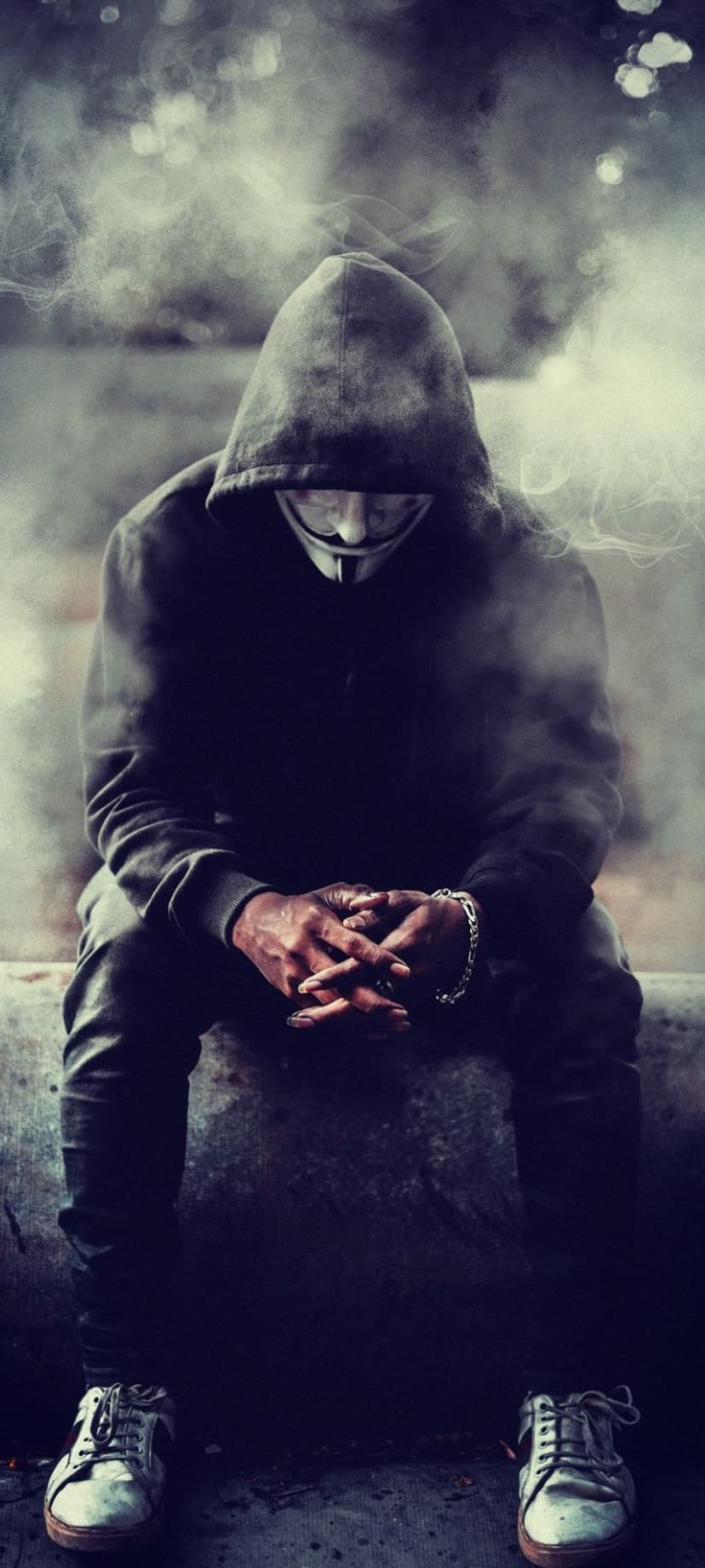 Guy Fawkes masked person in a black hoodie sits hunched over while surrounded by thick clouds of grey smoke.