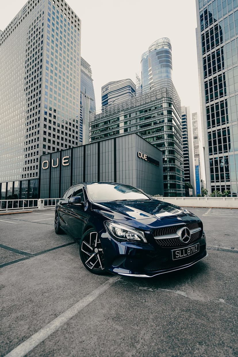 Mercedes Benz CLA with glowing headlights parked in a modern city plaza surrounded by glass skyscrapers at night.