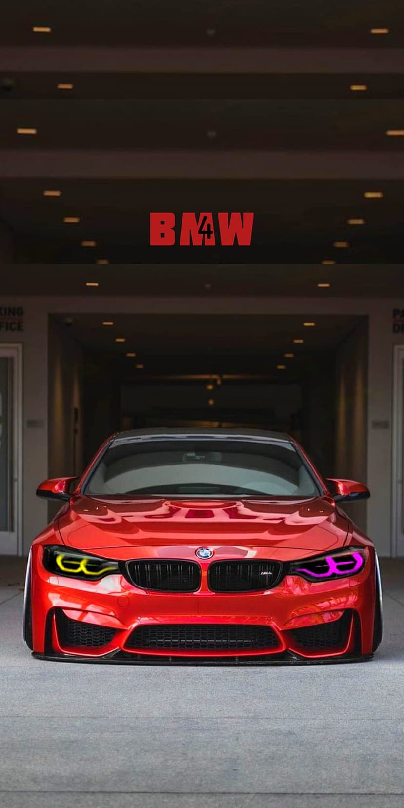 Red BMW M4 sports car with glowing yellow and pink headlights parked in a dark, contemporary garage setting.
