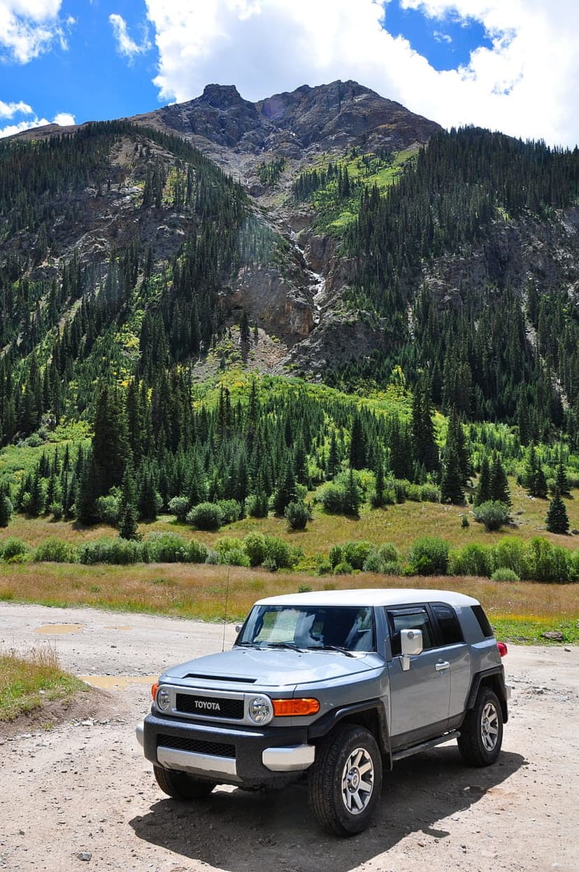 Silver Toyota FJ Cruiser parked on a dirt road with pine-covered mountains under a bright blue sky.