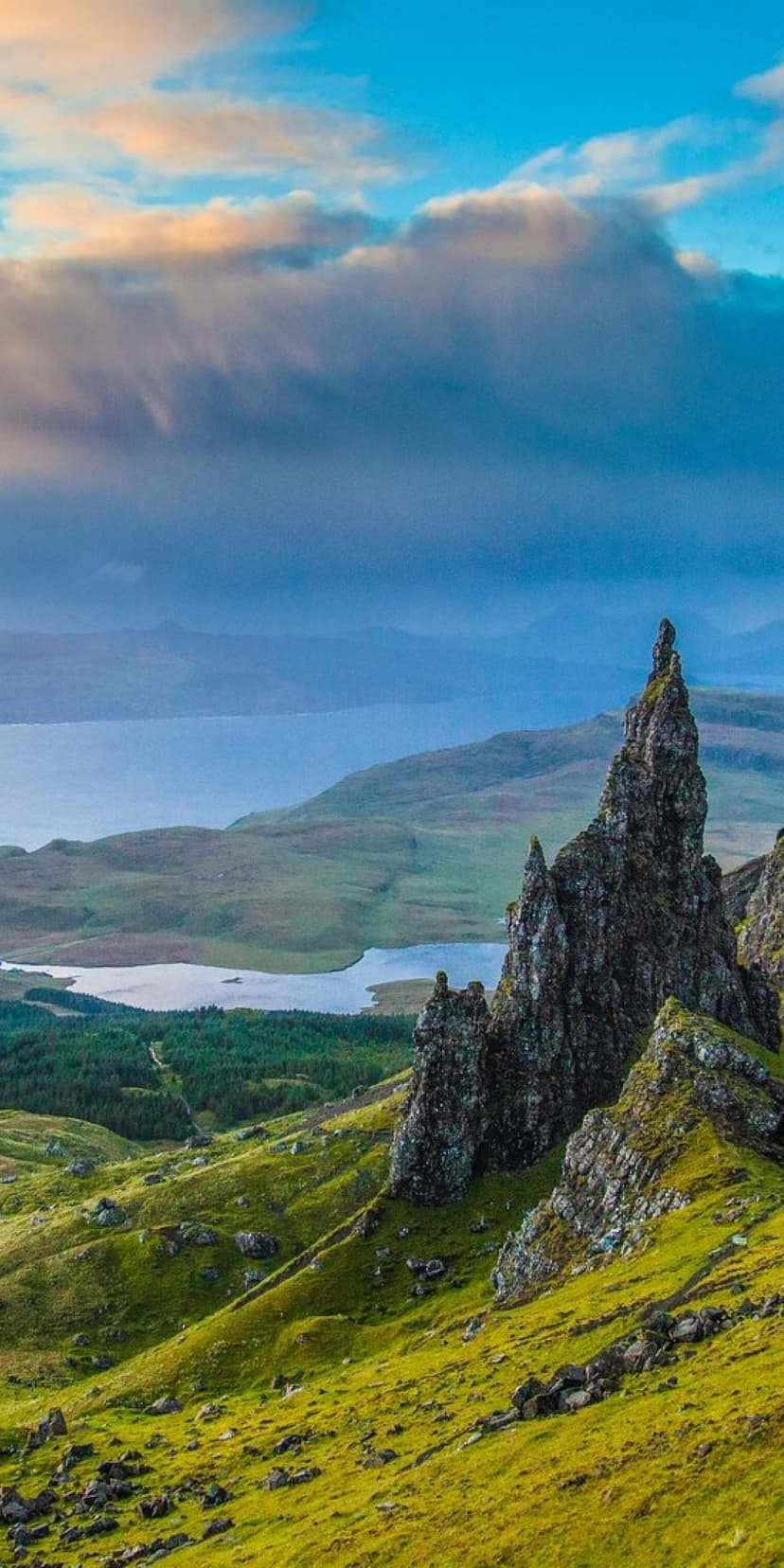 Old Man of Storr rock pinnacle stands above green hills and a calm lake under a cloudy morning sky in Scotland.