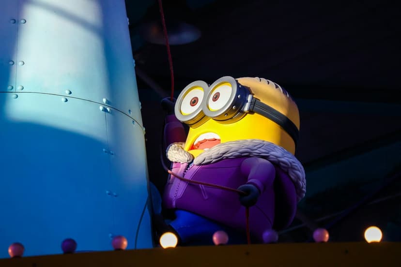 Minion wearing a purple winter jacket and goggles holds a red rope against a blue architectural background.