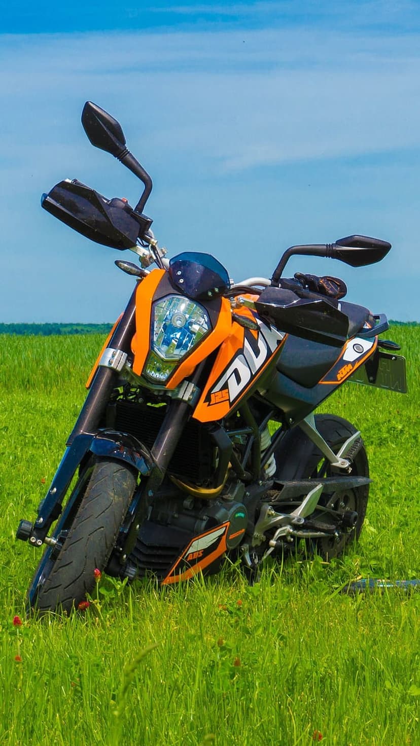 Orange KTM Duke motorcycle parked in a tall green field beneath a clear blue sky during a sunny afternoon.