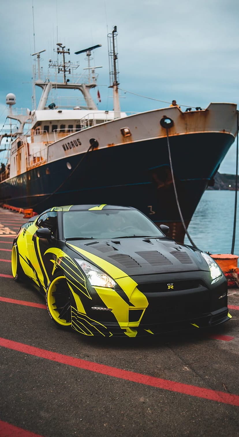 Nissan GT-R featuring neon yellow decals parked beside a massive cargo ship at an industrial dockside.
