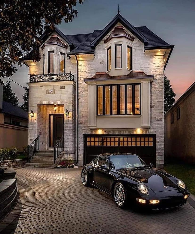 Black sports car parked on a stone driveway in front of a modern glass mansion illuminated at twilight.