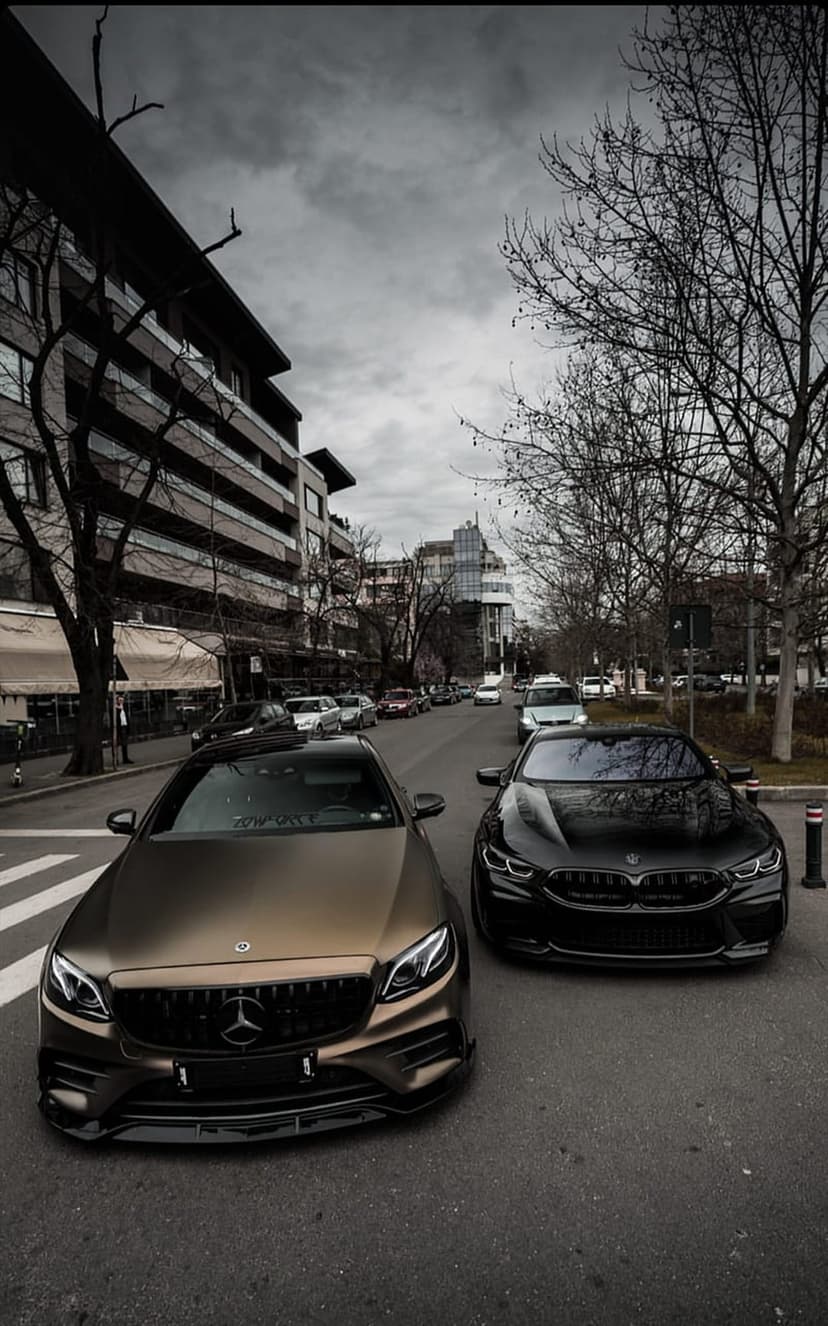 Bronze Mercedes and black BMW parked on a city street under a cloudy sky with leafless trees in the background.