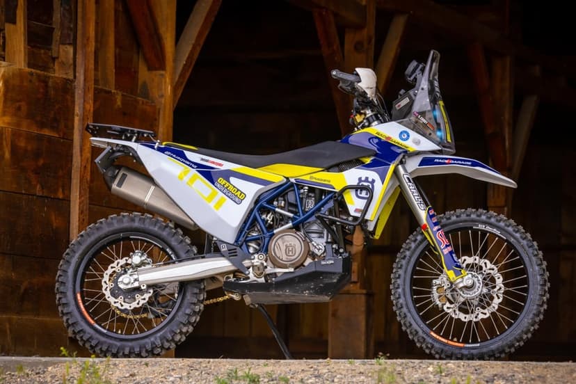 White Husqvarna 701 Enduro rally bike with blue frame and knobby tires parked in a dark wooden barn.