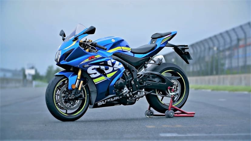 Blue Suzuki GSX-R sportbike on a red paddock stand over gray asphalt with a blurred race track background.
