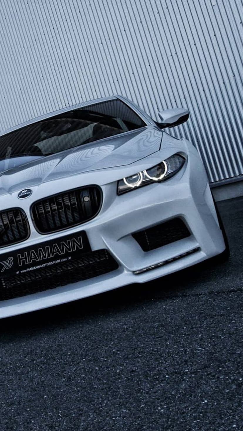 White BMW M5 with a Hamann front bumper and black grille parked against a grey corrugated metal wall on asphalt.