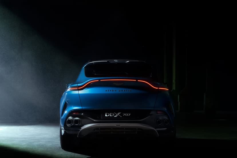 Blue Aston Martin DBX 707 rear view in a dark studio with glowing red taillights and dramatic spotlight beam.