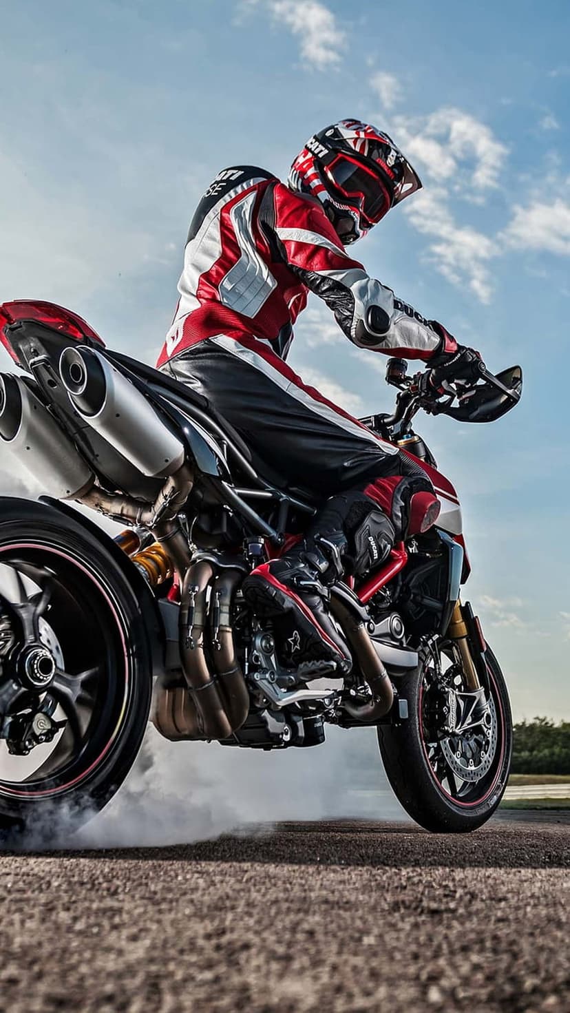 Motorcyclist in red racing gear leans into a smoky burnout on a sportbike against a cloudy gray sky backdrop.