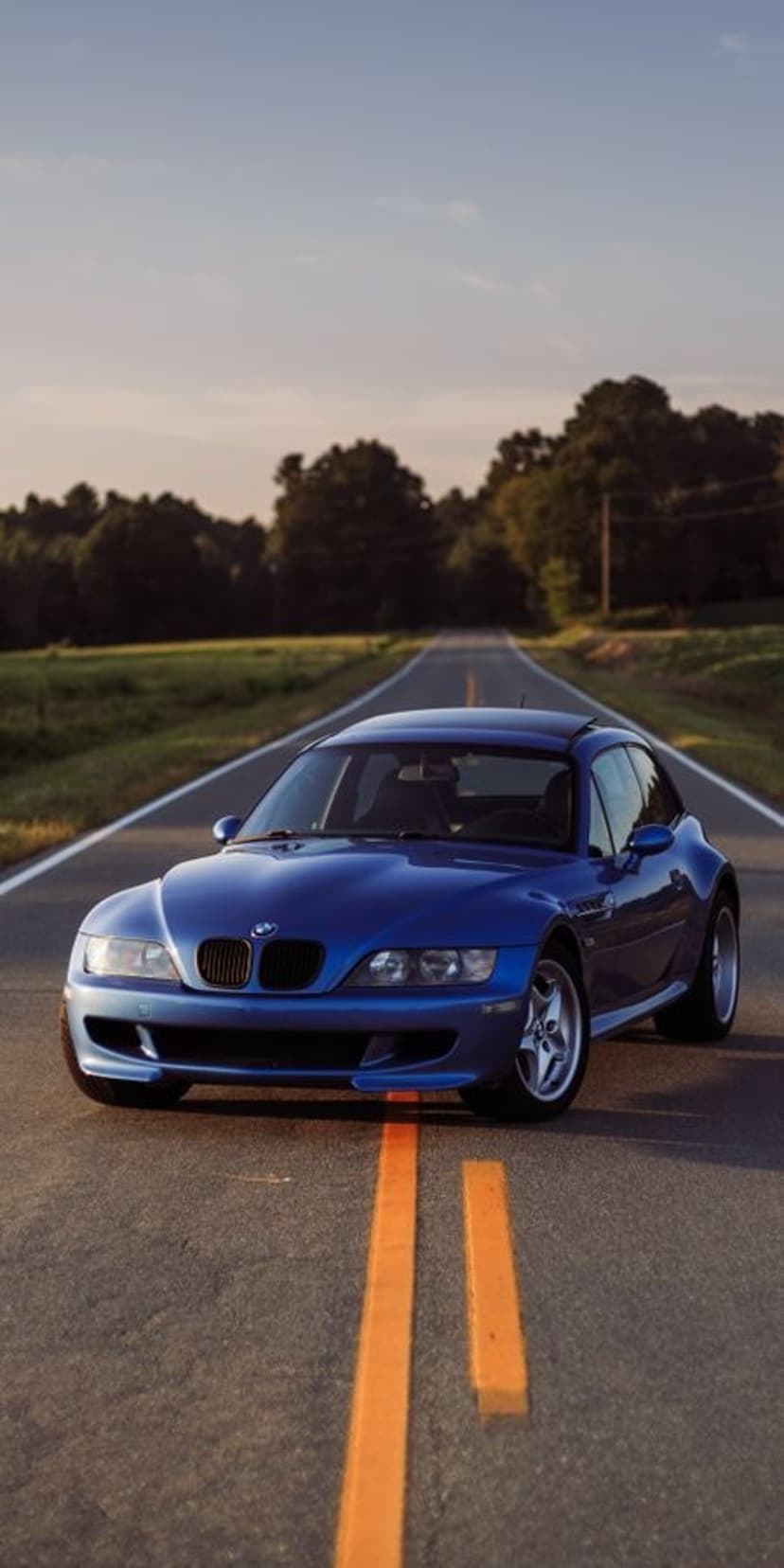 Blue BMW M Coupe driving on a two-lane country road between green fields under a hazy sunset sky at golden hour.