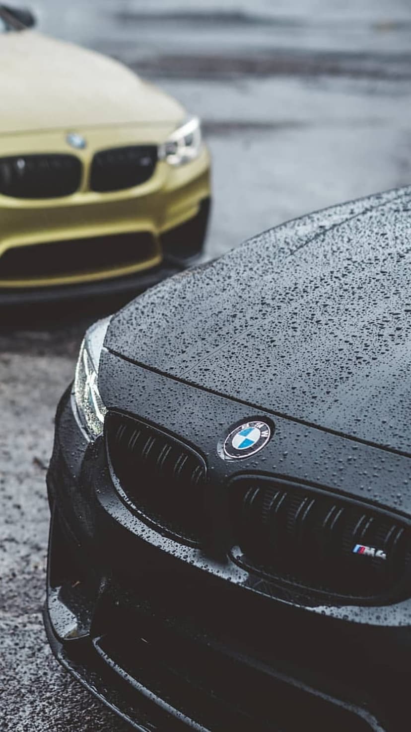 Black BMW M4 with raindrops on its hood parked in front of a blurred yellow car on a wet street.