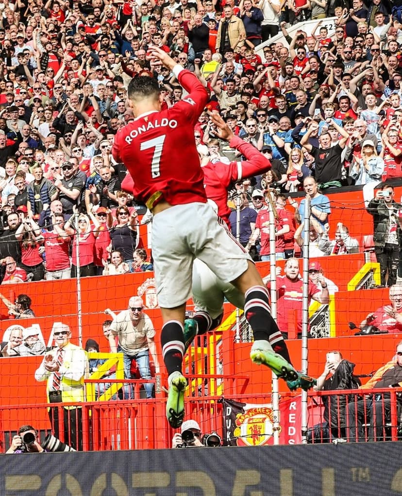 Cristiano Ronaldo in a red Manchester United kit celebrates a goal near cheering fans in a crowded stadium.