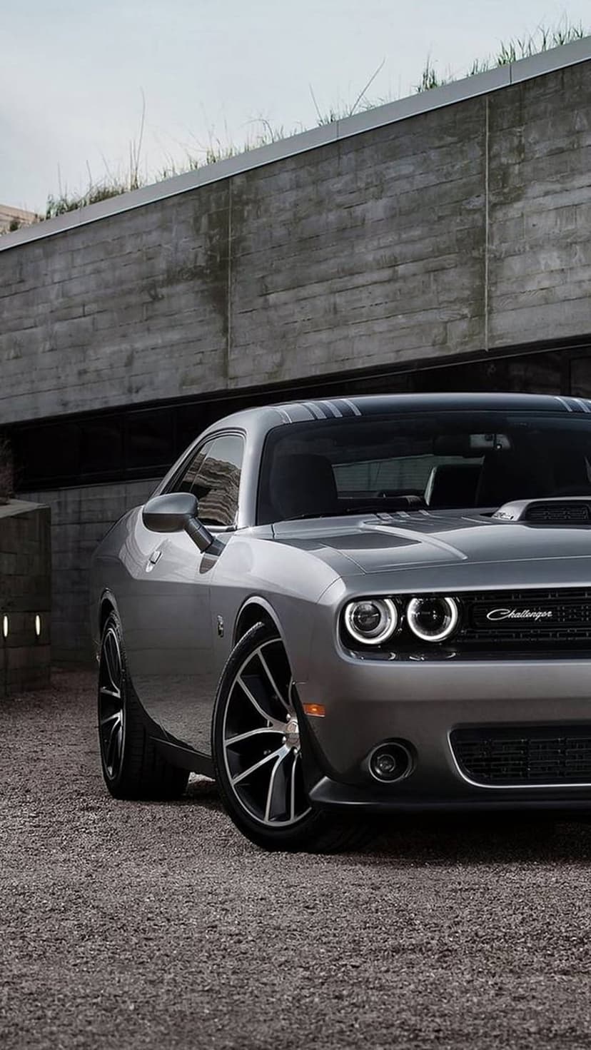 Silver Dodge Challenger Hellcat with black stripes parked on gravel in front of a modern concrete structure.