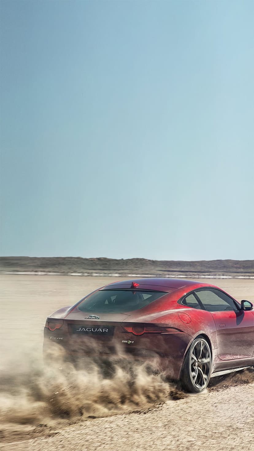 Red Jaguar F-Type sports car drifting on desert sand under a blue sky, kicking up a large cloud of golden dust.