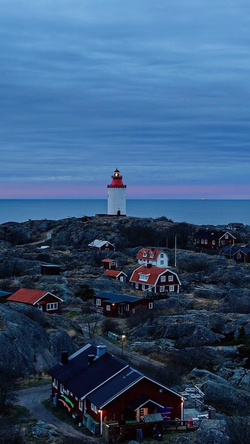 White lighthouse with a red lantern room overlooks small cottages and calm sea during a pink and blue twilight.