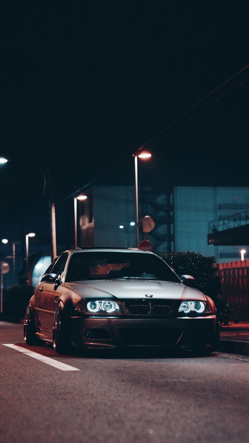 Silver BMW E46 coupe with custom headlights and lowered suspension parked on a dark city street at night.