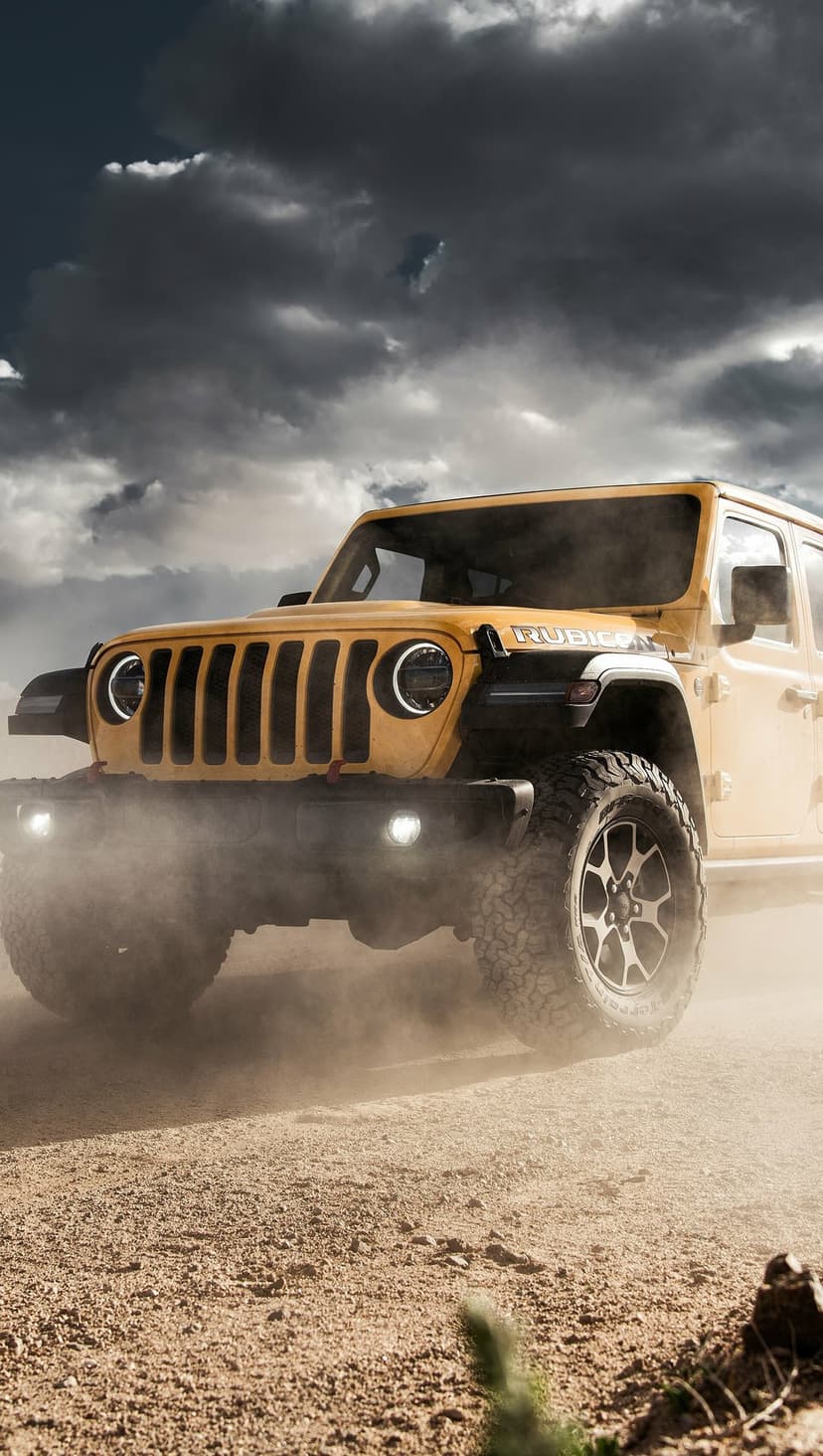 Yellow Jeep Rubicon kicking up dust on a desert trail under a dark stormy sky with glowing headlights.