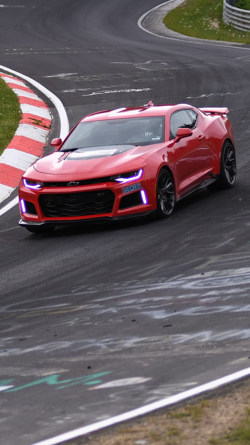 Red Chevrolet Camaro with purple headlights speeds around a wet track corner beside red and white curbing.