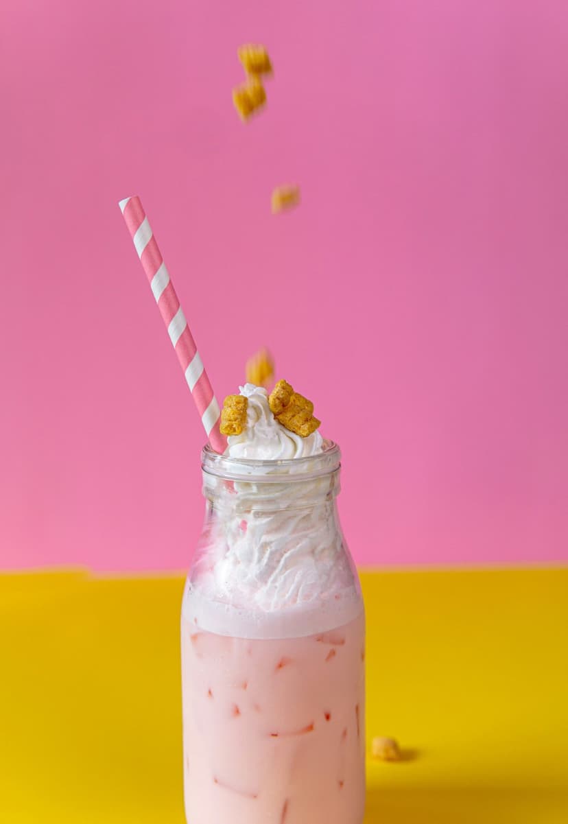 Strawberry milkshake in a glass bottle with whipped cream and cereal against a pink and yellow background.
