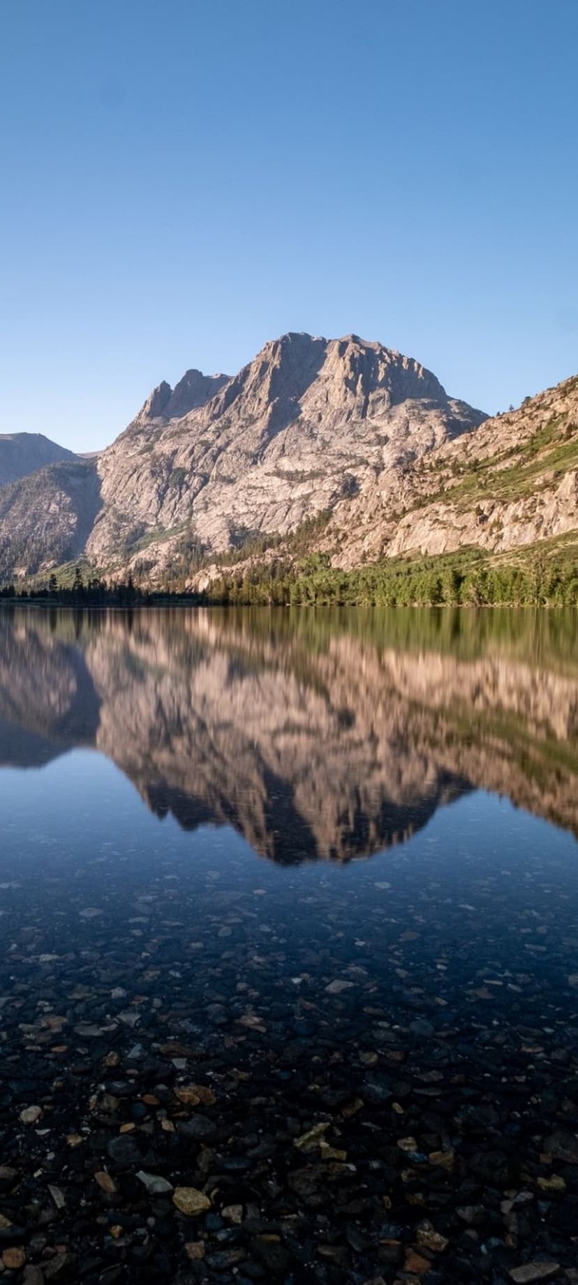 Jagged mountain peaks reflecting in a clear lake with colorful pebbles visible in the shallow water at sunrise.