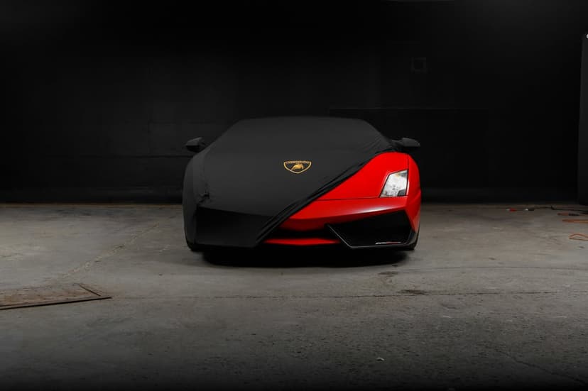 Red Lamborghini front bumper exposed under a black car cover with a gold emblem on a dark garage floor.