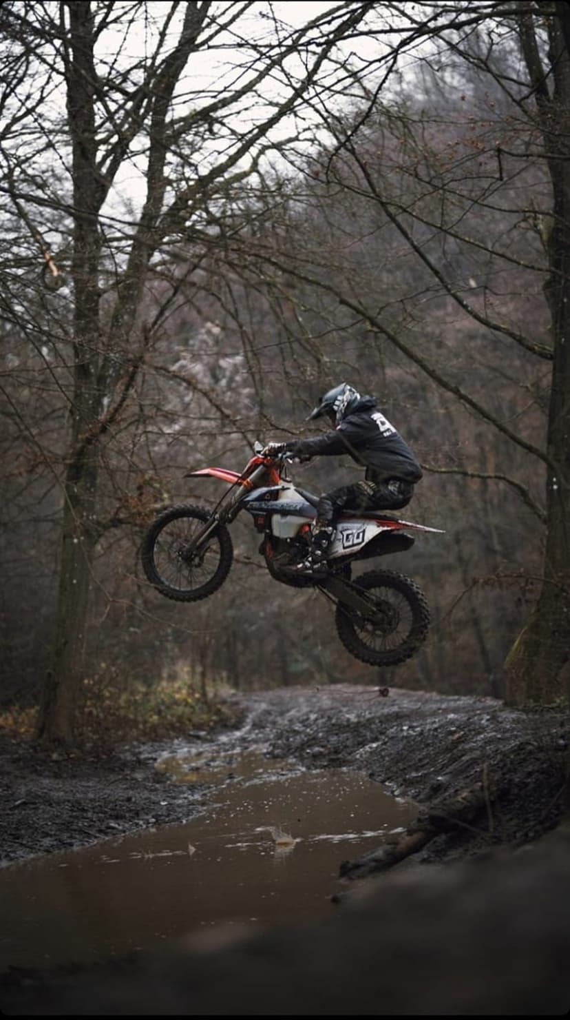 Motocross rider in a black hoodie jumps a dirt bike over a muddy puddle amidst bare trees in a dark forest.
