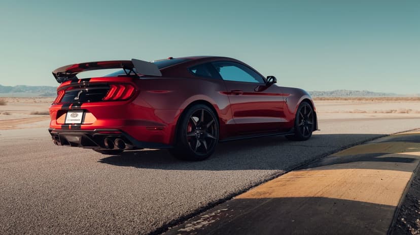 Red Ford Mustang Shelby GT500 with a large rear wing on a desert racetrack under a clear blue cinematic sky.