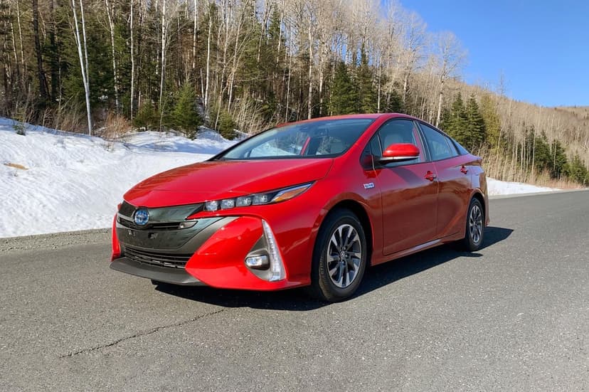 Red Toyota Prius Prime parked on an asphalt road with a snowy forest hillside and clear blue sky behind it.