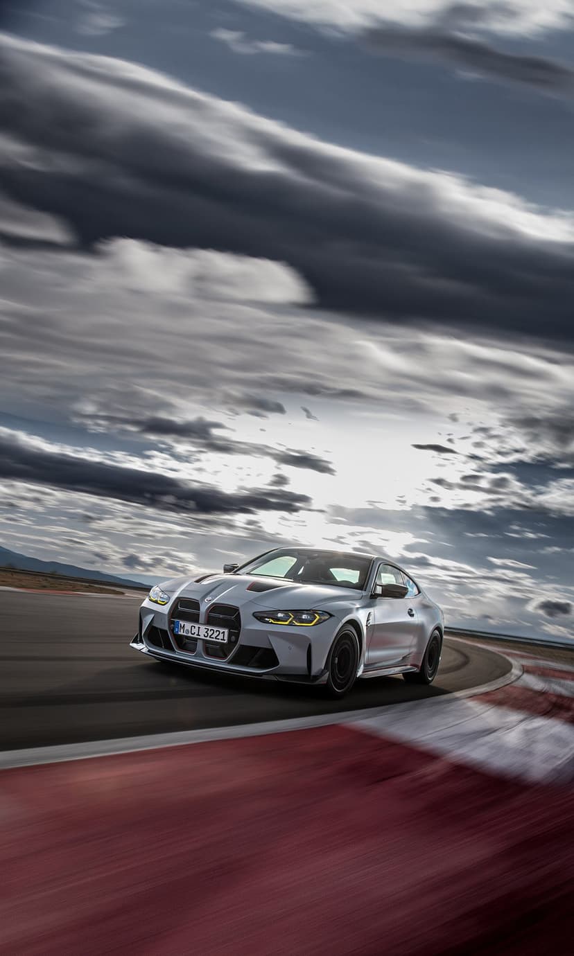Silver BMW M4 CSL with yellow headlights and red accents drifting on a racetrack under a cloudy sky.