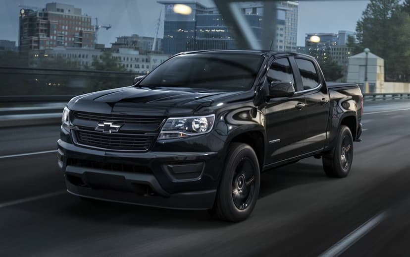 Black Chevrolet Colorado truck driving on a city bridge at dusk with glowing urban lights in the background.