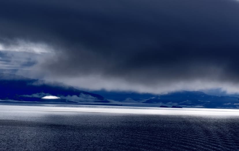 Heavy dark clouds hang above a rippling Arctic ocean with distant mountains and a thick layer of white sea mist.