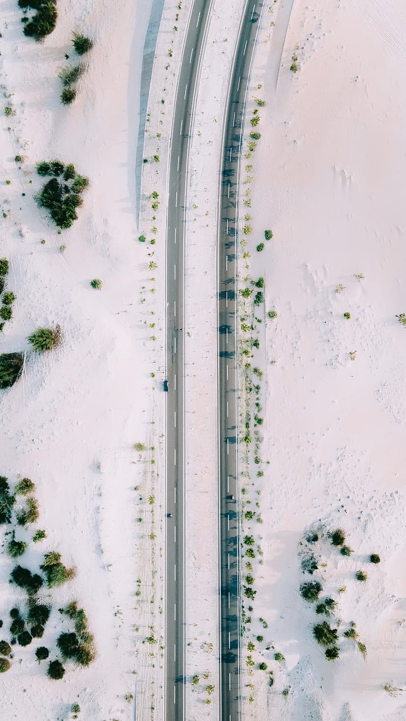 Dual highway carving through pale rippled sand dunes with sparse green shrubs from an aerial perspective.