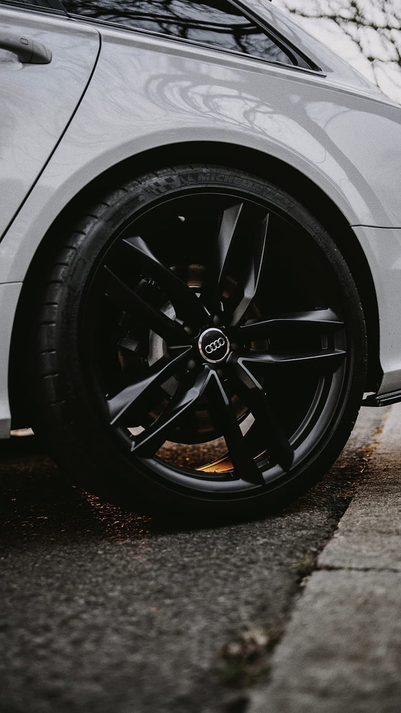 Black Audi alloy wheel with silver spokes on wet asphalt road featuring Michelin tire and metallic paint.