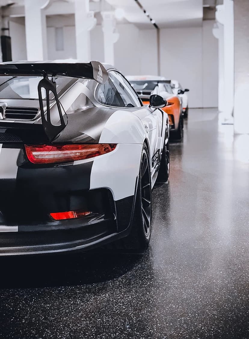 Porsche 911 with a black and white racing livery leads a row of sports cars reflecting on a polished showroom floor.