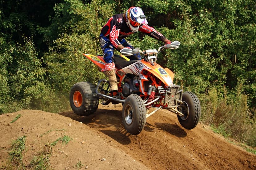 Motocross rider jumps an orange ATV over a dirt mound with a dense green forest background in clear daylight.