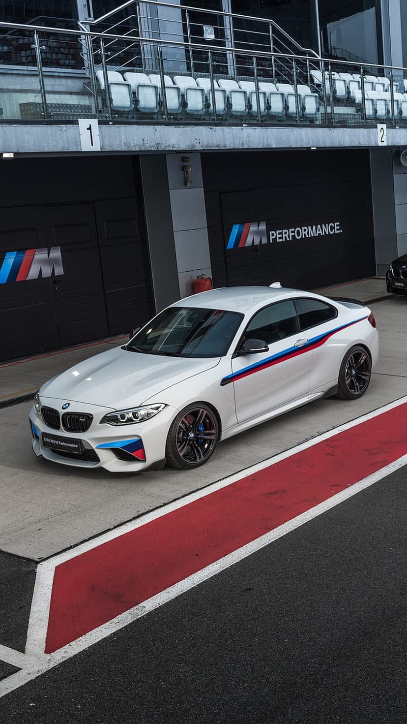 White BMW M Performance car with M stripes parked on a race track pit lane near red and white asphalt markings.