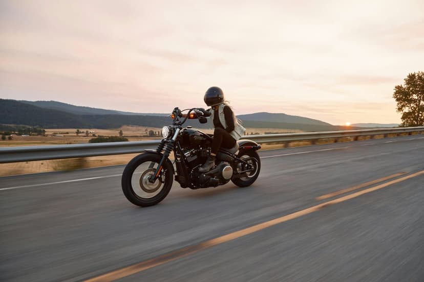 Motorcyclist riding a black bike on a highway toward a glowing orange sun and distant silhouetted mountains.