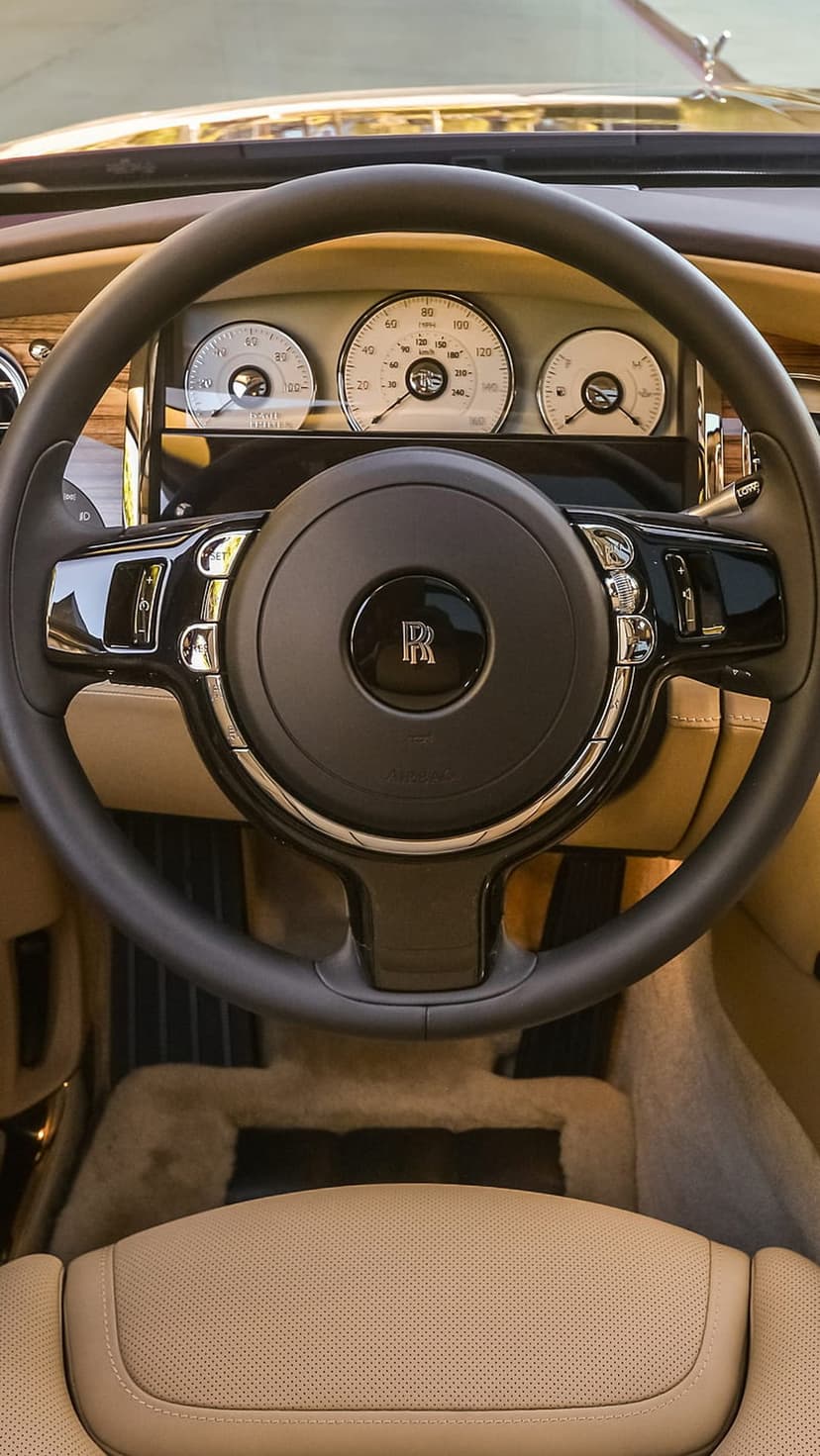 Rolls-Royce steering wheel with logo, tan leather seats, and chrome dashboard gauges in a luxury car interior.