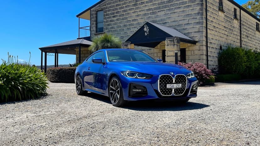 Blue BMW 4 Series Coupe parked on a gravel driveway in front of a limestone house under a bright blue sky.