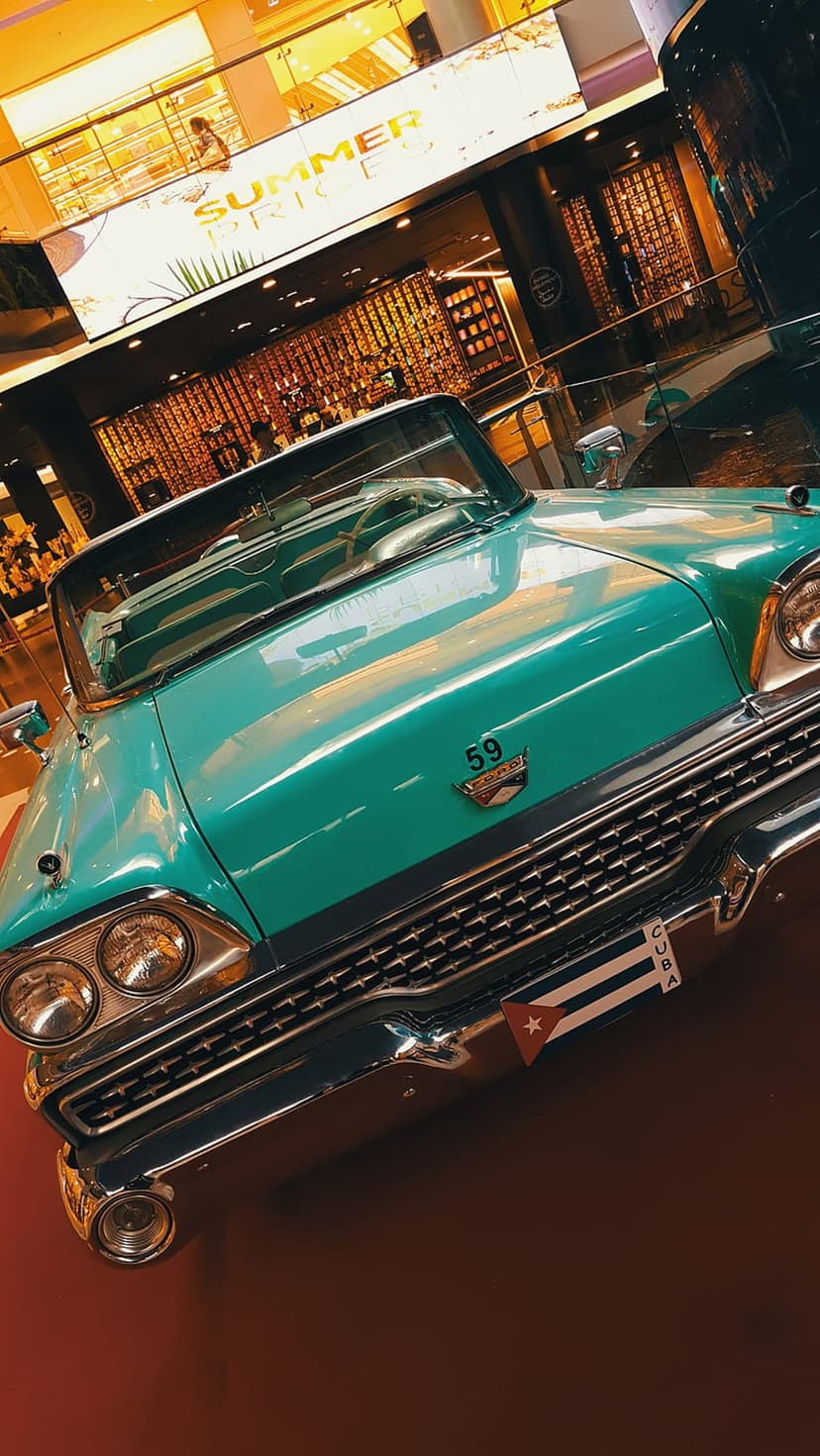 Teal 1950s convertible car with chrome grille and Cuba flag sticker on the bumper in a warm exhibition hall.
